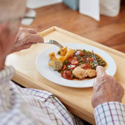 Une personne âgée est assise à une table, dégustant un repas composé de poulet, de pommes de terre, de petits pois et de carottes dans une assiette blanche - peut-être dans le cadre du repas domicile PACA - avec un verre d'eau à proximité, à l'aide d'un couteau et d'une fourchette.