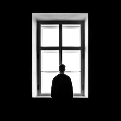 Une personne est assise, seule, en silhouette, face à une grande fenêtre éclairée par une lumière vive à l'extérieur, entourée d'obscurité.