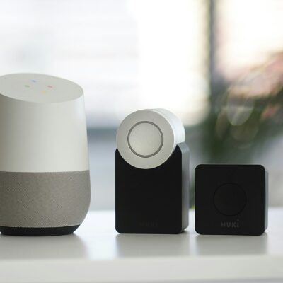 Une enceinte intelligente Google Home est posée sur une surface blanche à côté de deux serrures intelligentes Nuki noires, le tout sur un fond flou.