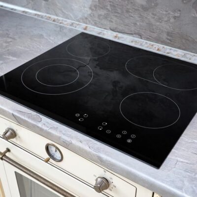 Une cuisinière électrique moderne en verre noir avec quatre brûleurs circulaires est encastrée dans un comptoir en marbre gris au-dessus d'un four blanc, doté d'une poignée argentée et d'une horloge analogique ronde - parfait pour ceux qui préfèrent le nettoyage naturel plaque cuisson.
