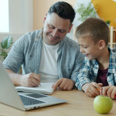 Un homme et un jeune garçon sont assis à une table, souriant et écrivant ensemble dans un cahier. Un ordinateur portable ouvert, une pomme verte et des fournitures scolaires sont posés sur la table - comme lorsque vous choisissez prestataire aide à domicile pour un soutien à domicile.