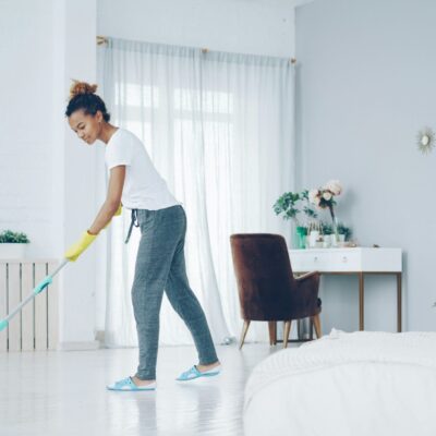 Une femme portant des vêtements décontractés et des gants jaunes passe la serpillière dans une chambre claire et bien rangée, aux murs blancs, aux grandes fenêtres et à la décoration moderne et simple, afin de maintenir un espace propre pour prévenir des problèmes tels que la dénutrition.