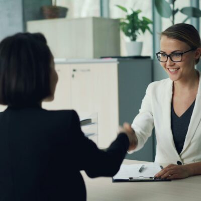 Deux femmes sont assises dans un bureau, souriantes et se serrant la main - peut-être en train de discuter du comparatif aide à domicile France. L'une porte un blazer blanc et des lunettes, l'autre une veste noire. Une planchette à pince et des documents sont posés sur le bureau.