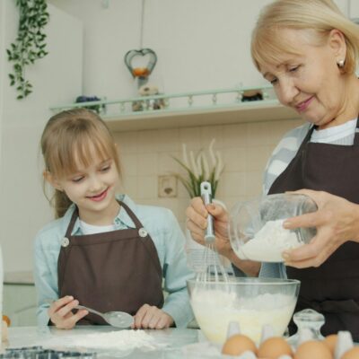 Une femme âgée souriante et une jeune fille, toutes deux vêtues d'un tablier, se tiennent dans une cuisine et mélangent des ingrédients dans un bol. Des œufs, du lait et des ustensiles de cuisine sont posés sur le comptoir pendant qu'elles cuisinent ensemble.